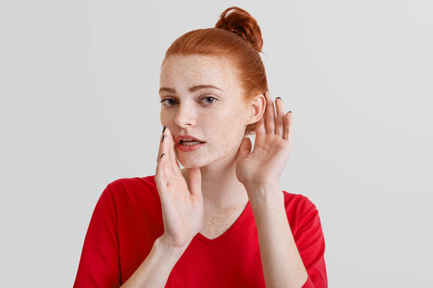 Beautiful foxy woman tries to overhear or eavesdrop someonen`s gossiping, keeps hand near ear, looks with secrecy, isolated over white background. Curious redhead young female listens something - Photo, image