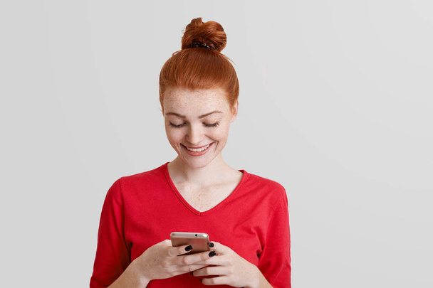 Attractive ginger young female in red clothes holds smart phone, types text messages, surfes social networks or online webpage, connected to wireless internet, isolated over white background - Foto, afbeelding