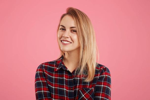 Pretty adorable woman with pleasant smile looks directly into camera, dressed in checkered shirt, isolated over pink background. Smiling friendly young female rejoices her success, poses indoor - 写真・画像