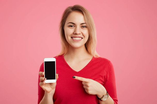 Alegre rubia sonriente hembra sostiene teléfono celular moderno con pantalla de copia en blanco, indica en ella, anuncia sus características, aislado sobre fondo de estudio rosa. Concepto de tecnología y personas
 - Foto, Imagen