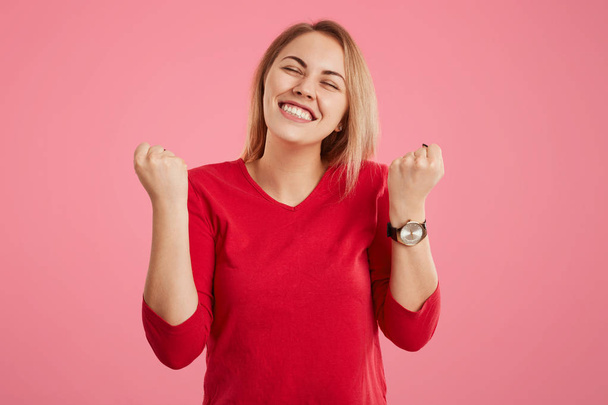 Glad female model clenches fists, dressed in red clothes, closes eyes with pleasure, dreams about something pleasant, isolated over pink background. Achievement, success and triumph concept. - Фото, изображение