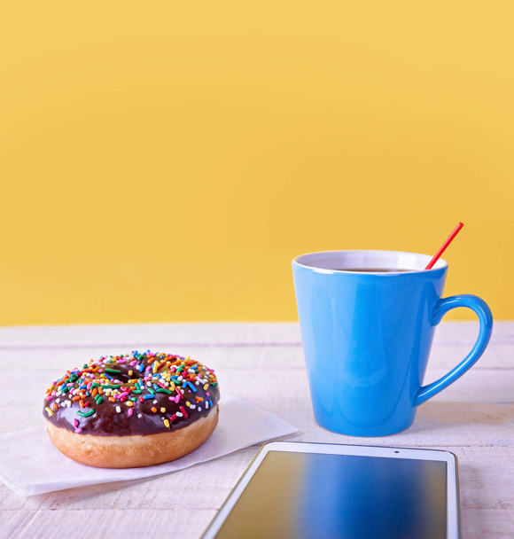 chocolate doughnut with colorful sprinkles on a napkin next to a blue coffee cup and a tablet on a wooden table with a yellow background - Фото, изображение