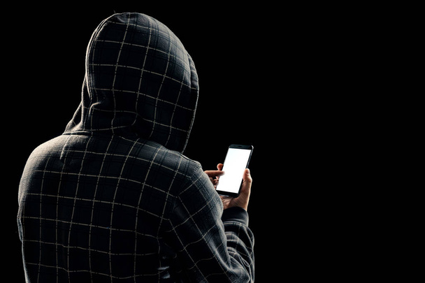 Silhouet van een man in een capuchon op een zwarte achtergrond, zijn gezicht niet zichtbaar is, de hacker houdt de telefoon in zijn handen. Het concept van een misdadiger, incognito, mysterie, geheimhouding, anonimiteit. - Foto, afbeelding