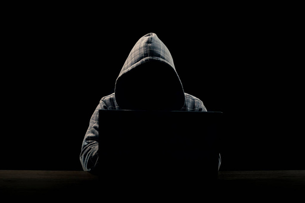 Silhouette eines Mannes mit Kapuze auf schwarzem Hintergrund, sein Gesicht ist nicht zu sehen, er sitzt am Computer. das Konzept eines Verbrechers, Inkognito, Mysteriums, Geheimhaltung, Anonymität. - Foto, Bild