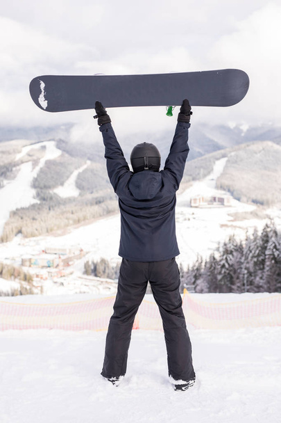 Ευτυχισμένος ο άνθρωπος στέκεται με snowboard πάνω από το κεφάλι του στην κορυφή του λόφου στα βουνά - Φωτογραφία, εικόνα