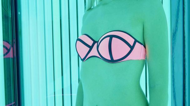 Crop image body of woman in pink and blue swimsuit standing in tanning booth - 写真・画像
