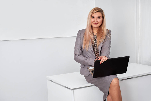 Ділова жінка в сірому костюмі, сидить на столі і працює на ноутбуці
 - Фото, зображення