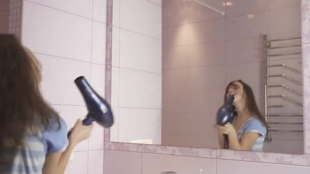 Szép boldog lány tinédzser szárítja a haját hajszárítóval és énekel és táncol, a fürdőszoba stock footage videóinak a tükör előtt - Felvétel, videó