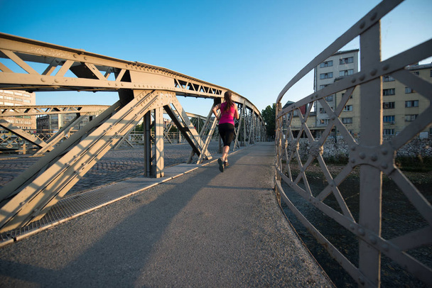 young sporty woman jogging across the bridge at sunny morning in the city - Фото, зображення