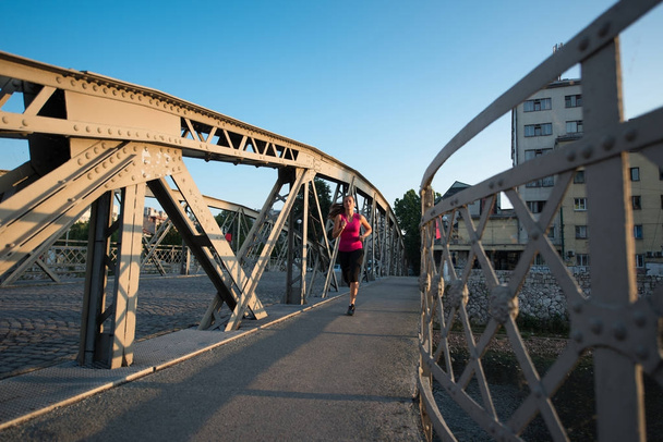 young sporty woman jogging across the bridge at sunny morning in the city - Photo, Image