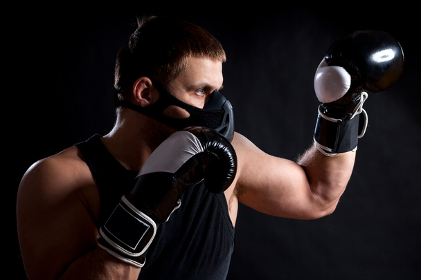 Jeune homme sportif aux yeux bleus en t-shirt noir sportif, portant des gants de boxe et un masque d'entraînement noir boxe sur un fond noir isolé
 - Photo, image