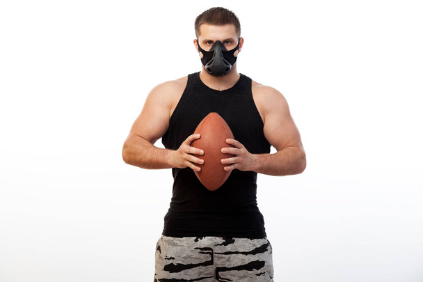 Young sporty blue-eyed man in a sporty black T-shirt, wearing boxing gloves and a black training mask is holding a rugby ball on a white isolated background - Fotografie, Obrázek