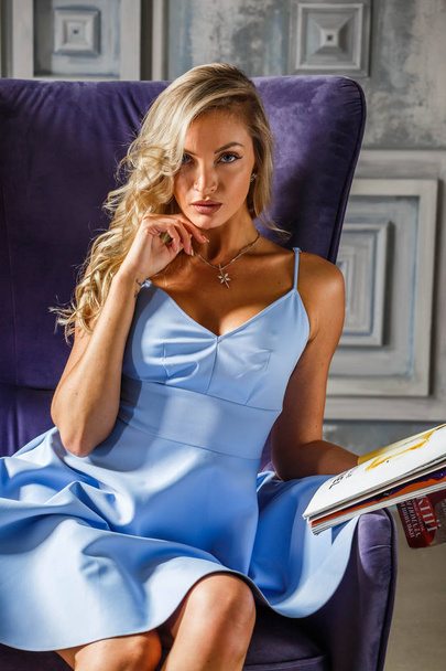 Studio portrait of beautiful blonde girl in blue dress. Sporty woman sitting in purple armchair. - Fotó, kép