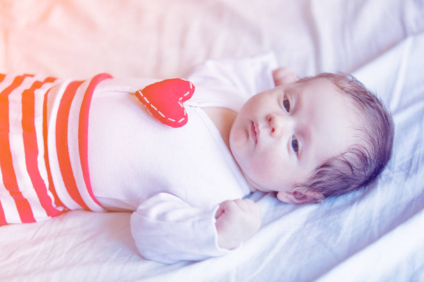 Pikkulapsi sydämen muotoinen lelu rinnassa makuulla valkoisella taustalla
 - Valokuva, kuva