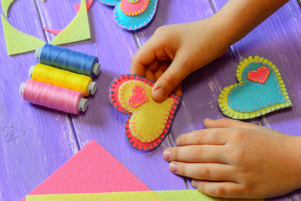 Kid holds a felt heart in his hand. Kid made a felt heart. Colorful felt hearts, scissors, thread on a wooden table. St. Valentines Day crafts concept. Simple hand sewing projects idea for children. Homemade Valentines Day gift idea. Heart crafts - Photo, Image