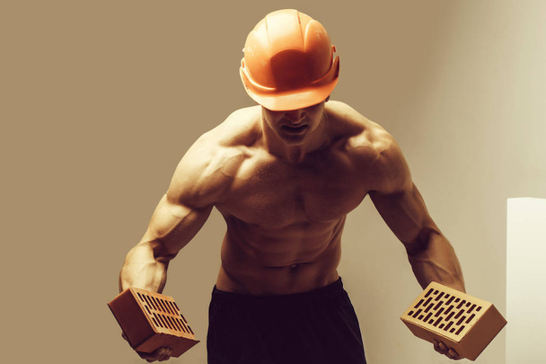 young handsome macho man builder with sexy muscular athletic strong body has bare torso and strong belly with six packs or abs in orange hard hat or helmet holds brick - Valokuva, kuva