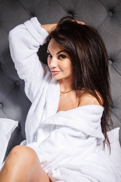 Studio portrait of beauty young brunette woman with professional makeup wearing white  bathrobe. Beautiful woman sitting on bed with grey headboard - 写真・画像