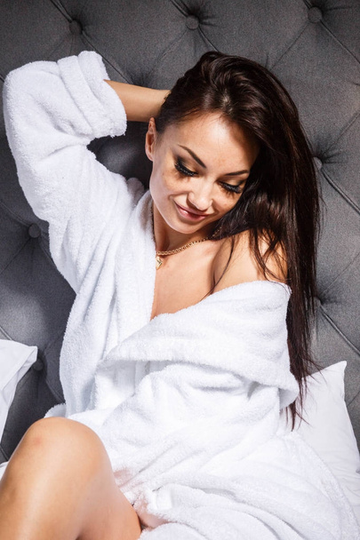 Studio portrait of beauty young brunette woman with professional makeup wearing white  bathrobe. Beautiful woman sitting on bed with grey headboard - Фото, изображение