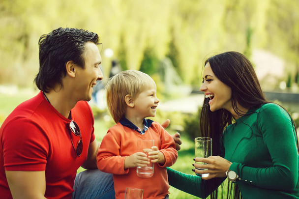 Happy family couple of young pretty woman and handsome man in park with son holding glasses of water and smiling sunny day - 写真・画像