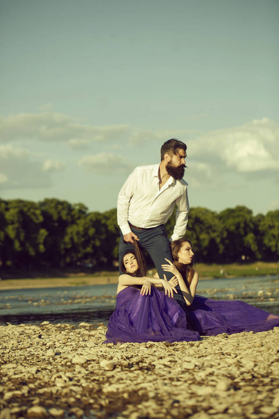 Bearded handsome man in white shirt standing with two young pretty women with long hair in violet dresses embracing male legs near water sunny day outdoor on blue sky background - Foto, Bild