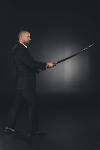 side view of mature man in suit with katana sword on dark background - Фото, зображення