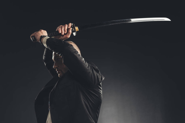 vista lateral del hombre en traje haciendo golpe con espada katana aislado en negro
 - Foto, imagen