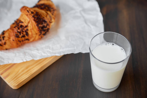 Tuore croissant valkoisella rypistyneellä paperilla ja puulauta lasillisella maitoa. Tuoretta croissantia tummalla puupöydällä
. - Valokuva, kuva