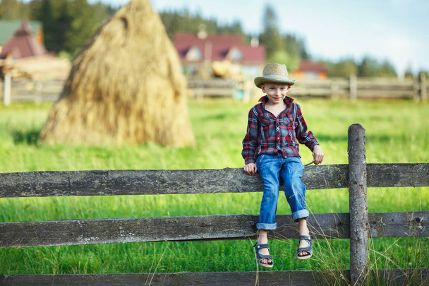 Little boy in hat sitting on fence against of haystack. Child on a hedge against backdrop of rural landscape. Young cowboy smiling shyly and looking at camera. Village, country trip, rest, concept - Foto, afbeelding
