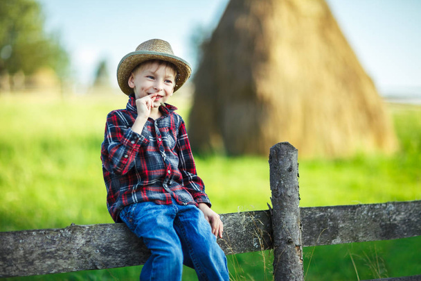 hild sits on wooden fence with straw in mouth against picturesque haystack, close up. Young boy in casual clothes resting on fence in village. Happy childhood concept. Vacation - Foto, Imagen