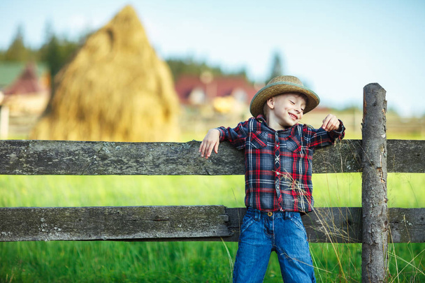 Smiling little boy leaned his back on wooden fence. Happy childhood in village landscape - Фото, зображення