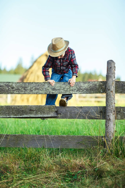 Small cowboy climbs over wooden fence, outdoor. Boy plays outdoors on wooden hedge with haystacks backdrop. Little traveler explores new opportunities - Foto, Imagem