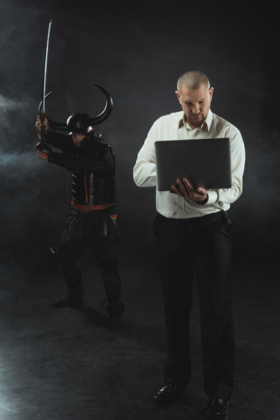 Konzentrierter Mann mit Laptop, während Samurai mit Schwert hinter ihm steht - Foto, Bild