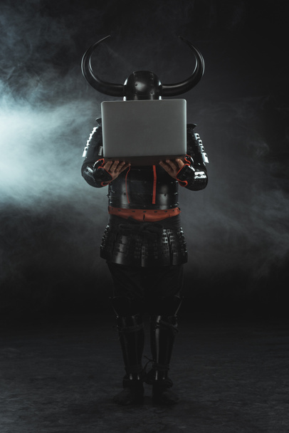 armored samurai holding laptop on dark background with smoke - Foto, Imagem