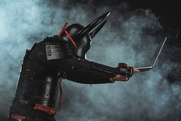 vista lateral de samuráis blindados sosteniendo portátil sobre fondo oscuro con humo
 - Foto, Imagen