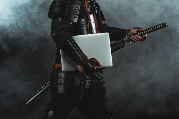 cropped shot of samurai in traditional armor with laptop taking out sword on dark background with smoke - Fotó, kép