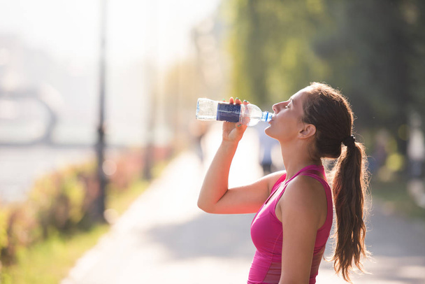 Athlete woman drinking water from a bottle after jogging in the city on a sunny day - 写真・画像