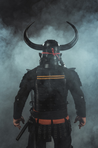back view of samurai in traditional armor and horned helmet on dark background with smoke - Фото, зображення