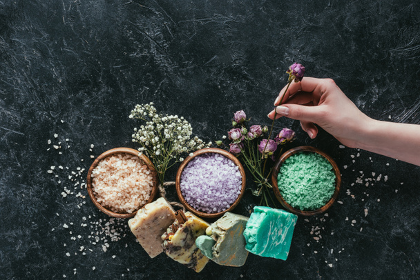 cropped view of female hand with natural homemade soap, dried flowers and sea salt for spa on black marble surface - Fotografie, Obrázek