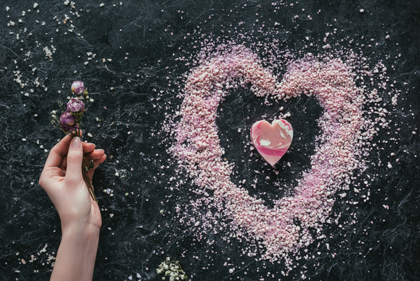 cropped view of female hand with dried roses on black marble surface with pink heart shaped soap and sea salt for spa on valentines day - Photo, Image