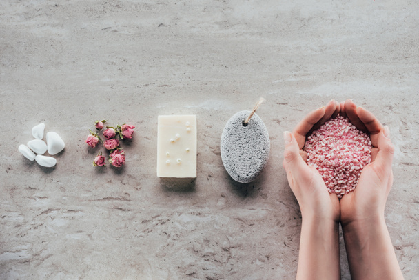 cropped view of hands with sea salt on marble surface with stones, dried roses, natural soap and pumice for spa - Фото, зображення