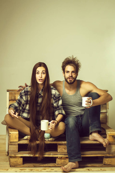 Young surprised couple of pretty girl or cute woman, with brunette long hair and handsome man or muscular macho with muscular torso, sits on wooden with tea cups, pallet sofa on grey background - Photo, image