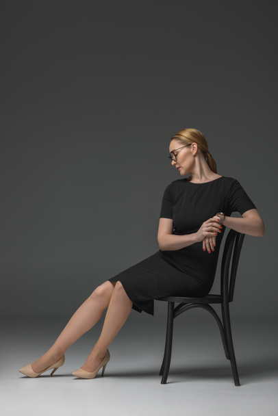full length view of stylish kazakh woman in eyeglasses and fashionable black dress sitting on chair on grey - Foto, afbeelding