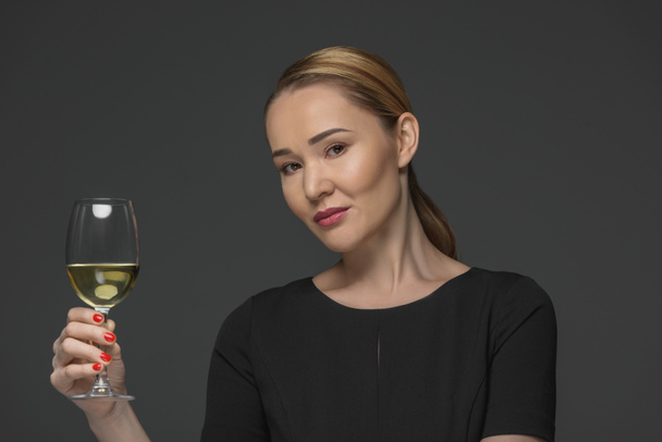 smiling kazakh woman holding glass of wine and looking at camera isolated on grey - Foto, immagini