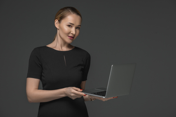 beautiful kazakh woman holding laptop and smiling at camera isolated on grey - Foto, afbeelding