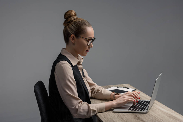 side view of kazakh businesswoman in eyeglasses using laptop at table isolated on grey  - Fotografie, Obrázek