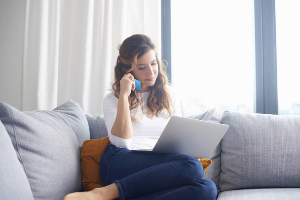 Mature woman sitting on sofa and looking thoughtful while shopping online with her credit card. - Photo, image