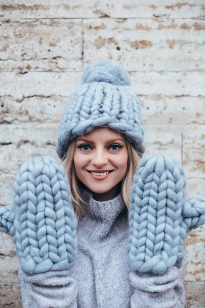 Young blonde woman in smoky blue knitted hat, knitted mittens and blue jeans make posing with stone grey wall on the background - Foto, afbeelding