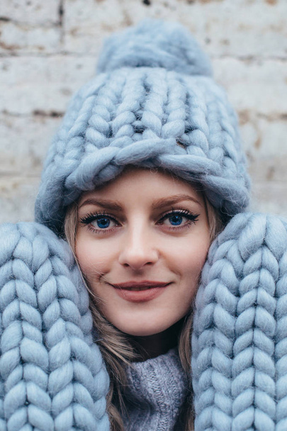 Young blonde woman in smoky blue knitted hat, knitted mittens and blue jeans make posing with stone grey wall on the background - Фото, изображение