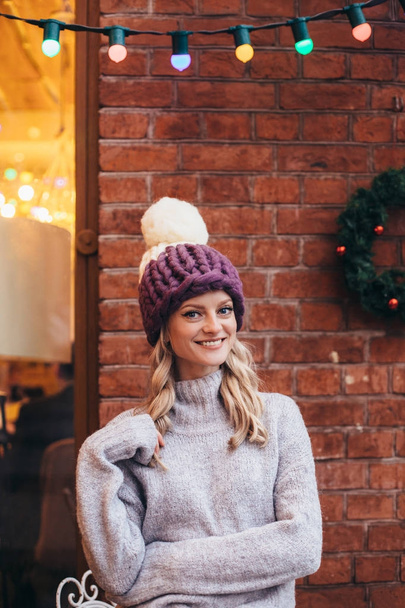 Blonde woman in purple knitted hats and grey sweater make posing with brick wall and Christmas garland on the background - 写真・画像