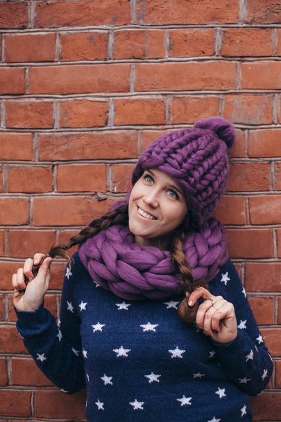Brunette woman in purple knitted hat, knitted infinity scarf and blue sweater with white stars on it make posing with brick wall on the background - Foto, afbeelding